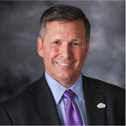 Greg's headshot. Man smiling wearing black suit with blue shirt and purple tie